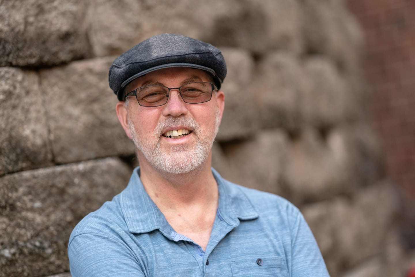 Randy Hatfield, dressed in a blue collared shirt and wearing glasses and a black hat, smiling outside.