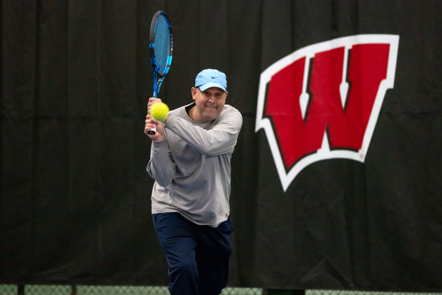 Andy swinging a tennis racket in front of a backdrop with the Wisconsin Badgers' motion W logo on it.