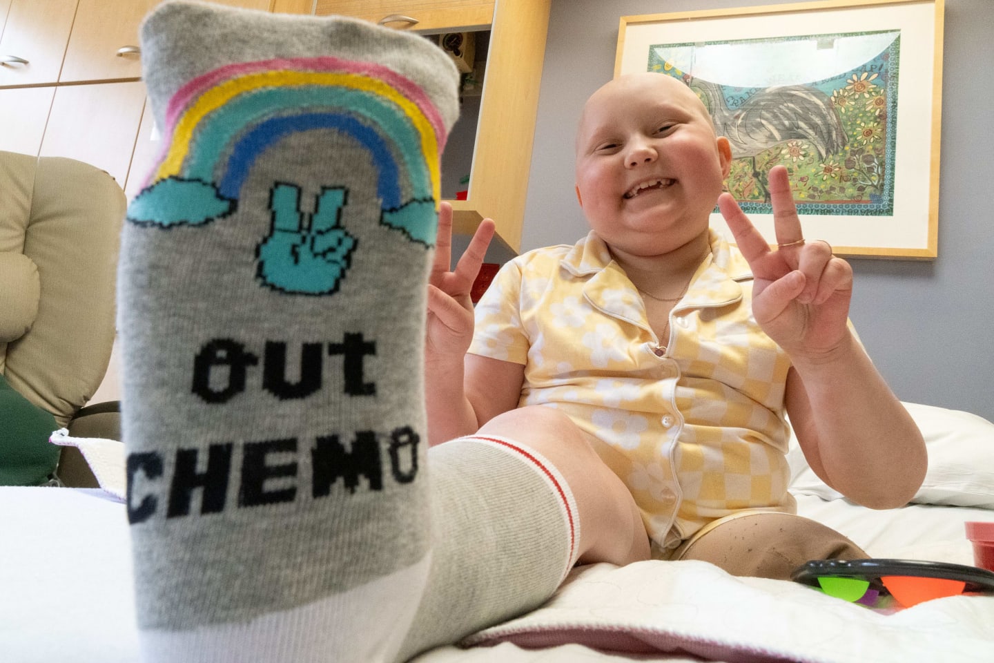 Girl sitting up in bed, smiling, with bottom of foot in foreground, with a sock on that says, "Peace out, chemo."