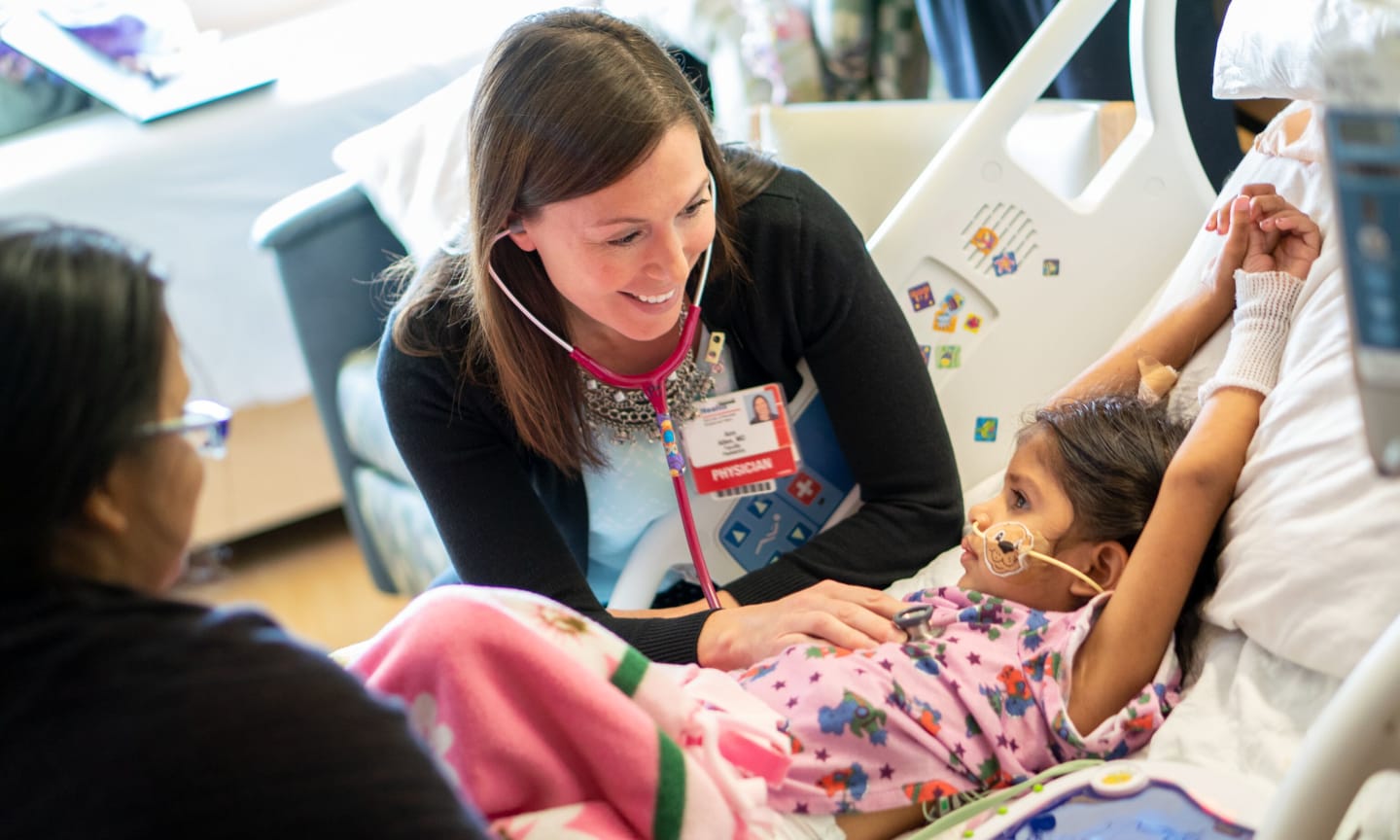 A female physician using a stethoscope on a little girl lying in a hospital bed