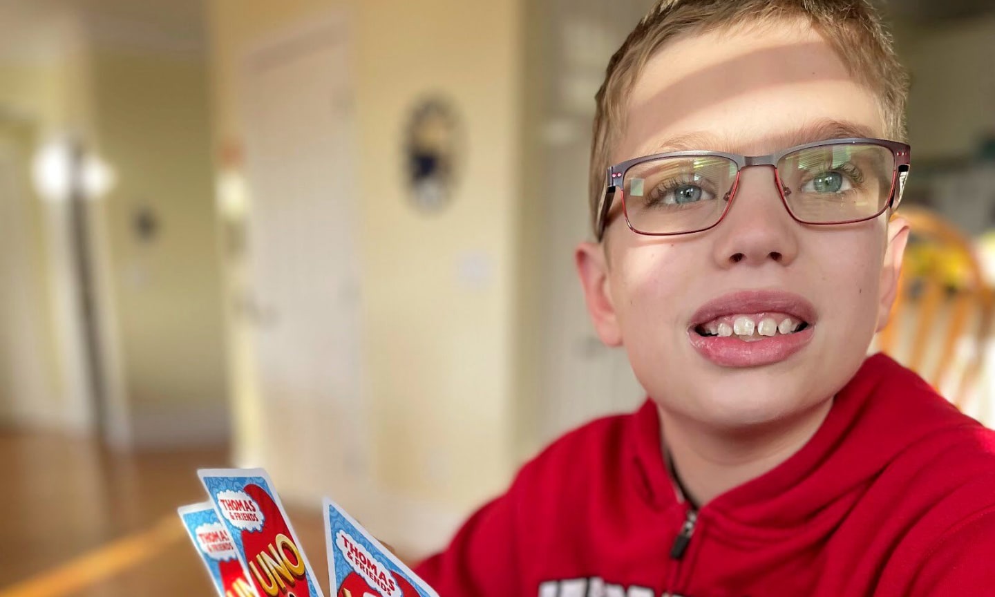 Luka, about 11 years old, smiling in a red sweatshirt and glasses while sitting at a table playing Uno.