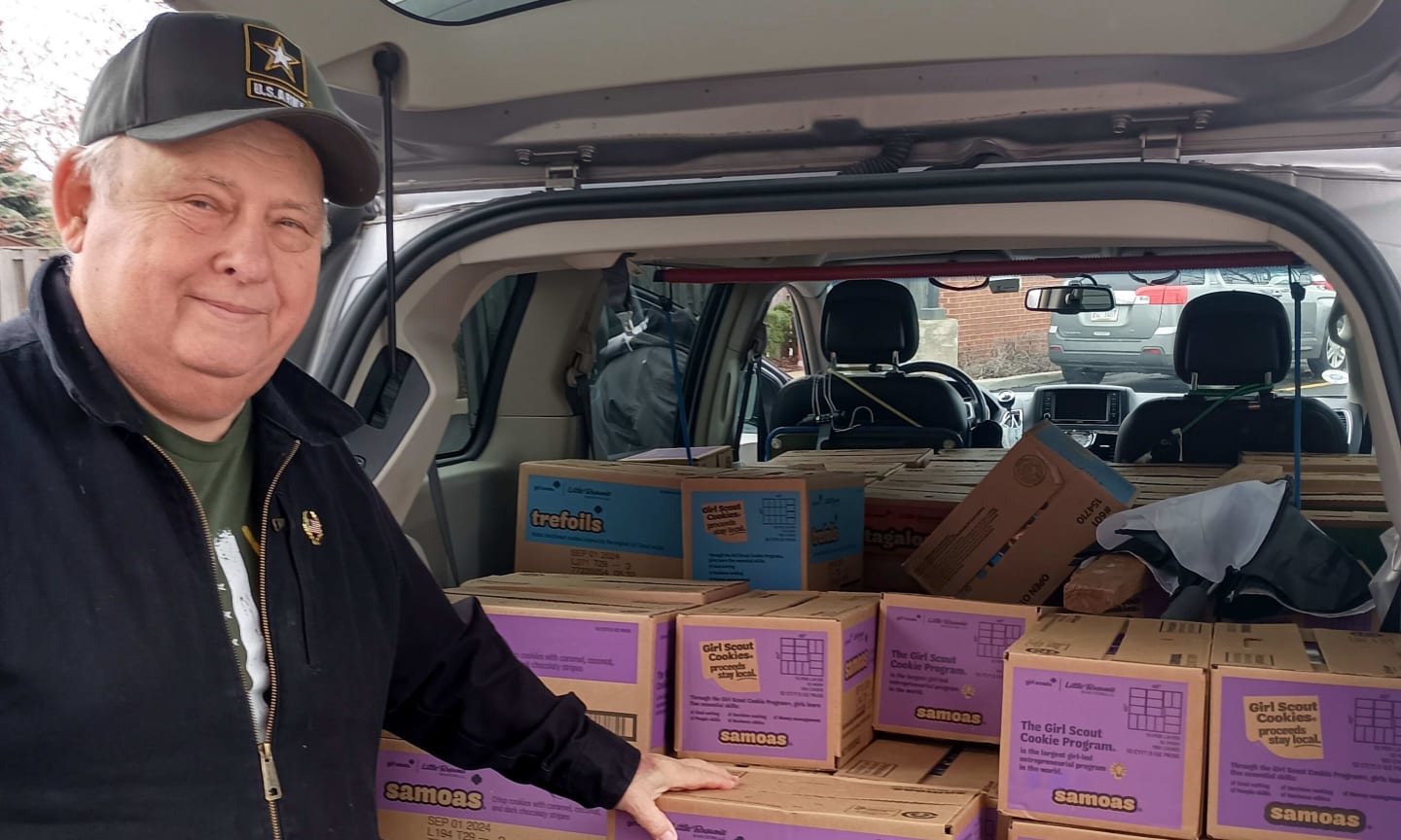 A man in a cap smiling and standing at the trunk of a car filled with boxes of cookies.