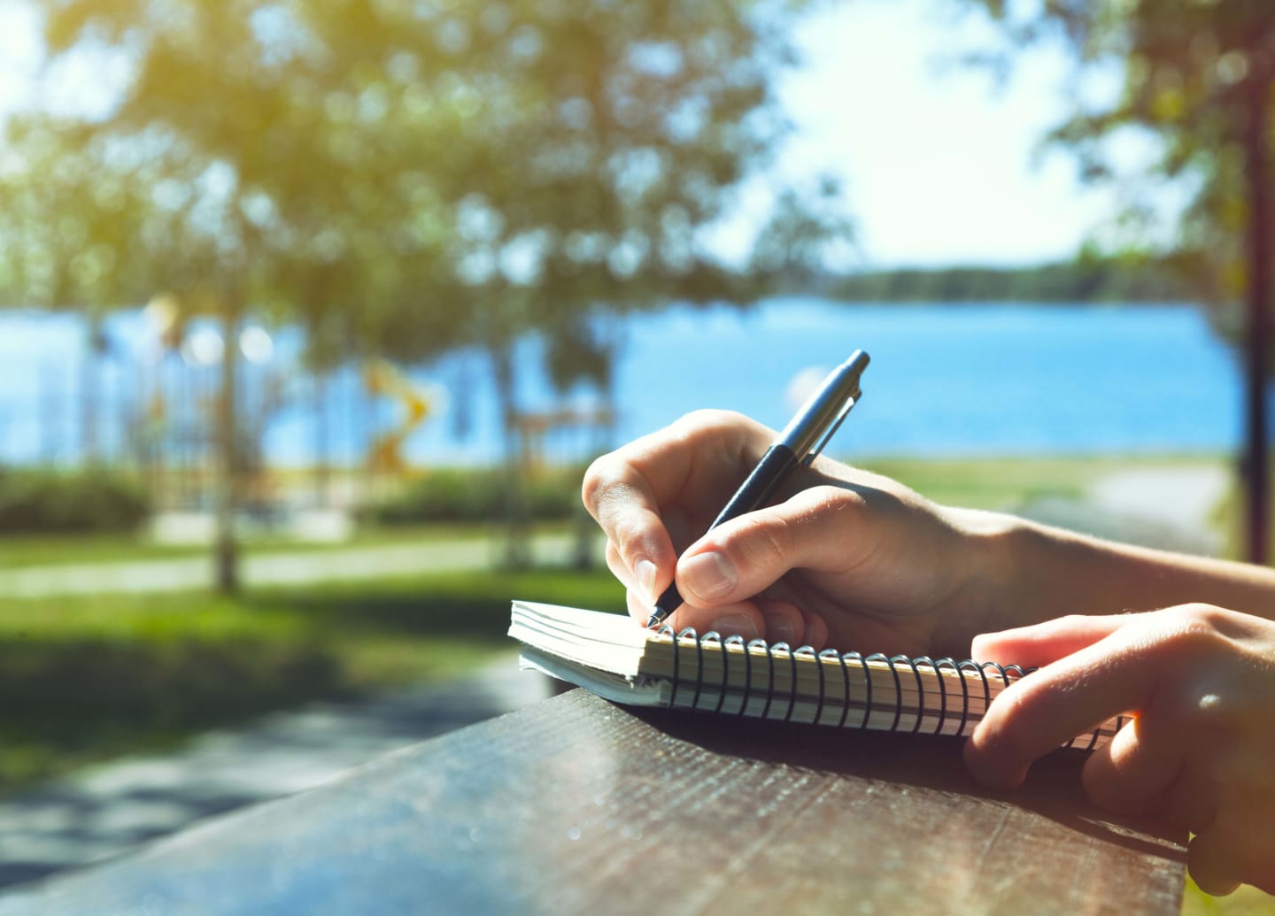 close up of hand holding a pen and writing in a notebook with a lake and playground in the background