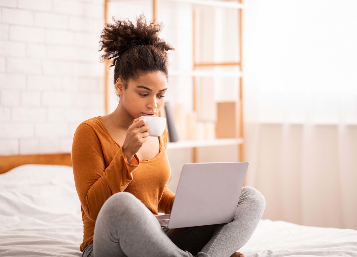 Individual sitting on bed looking at open laptop while drinking tea