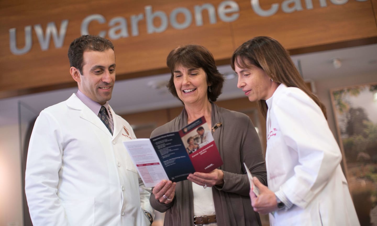 Jane DeShaw reading a pamphlet next to two doctors in front of a UW Carbon Cancer Center sign