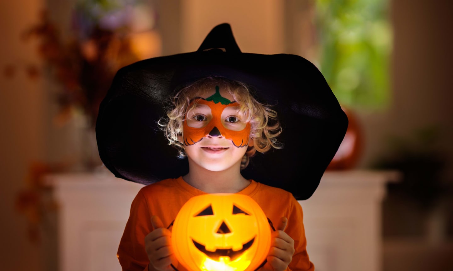 Child in a witch hat and orange shirt holding a glowing pumpkin