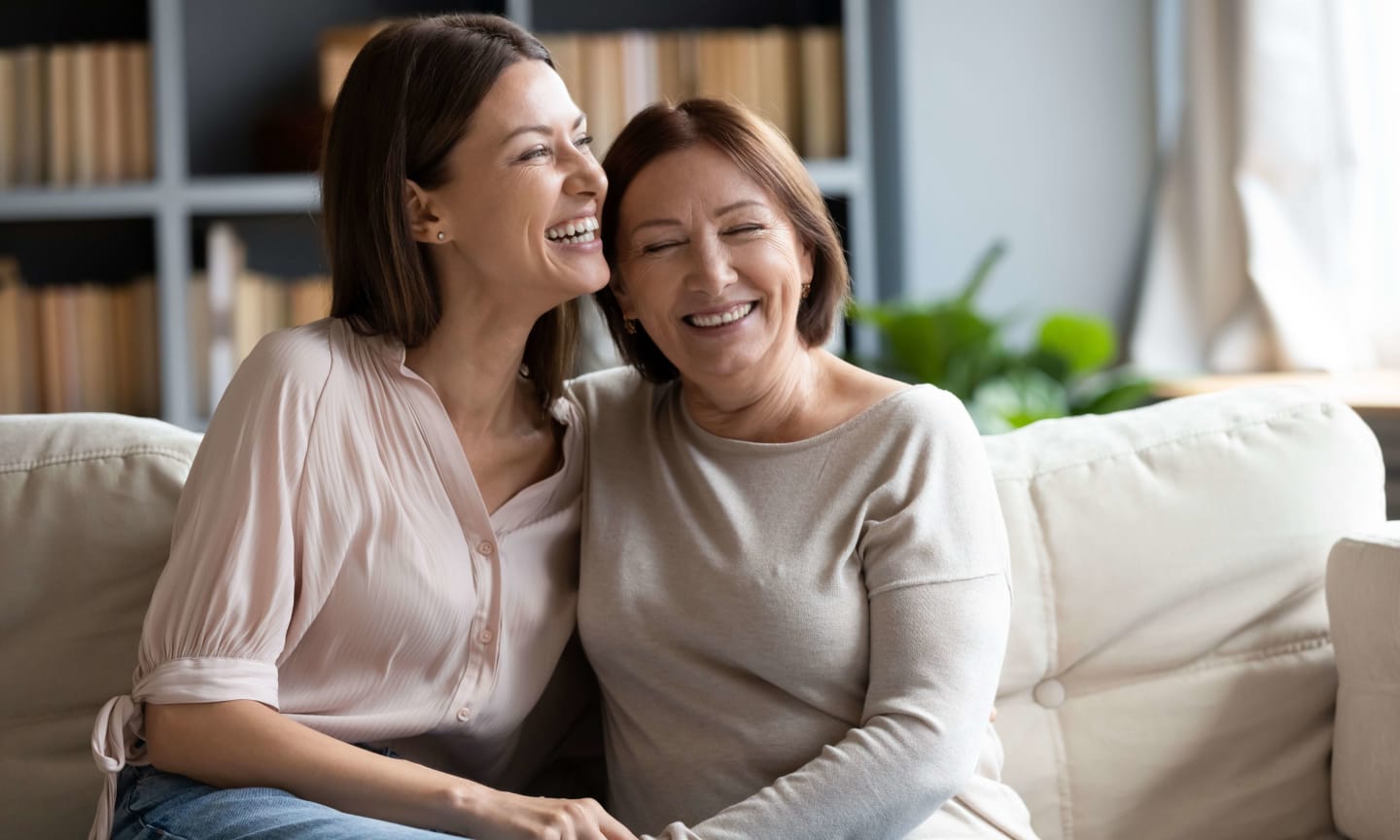 Adult daughter and her mother sitting on sofa with arms around each other laughing