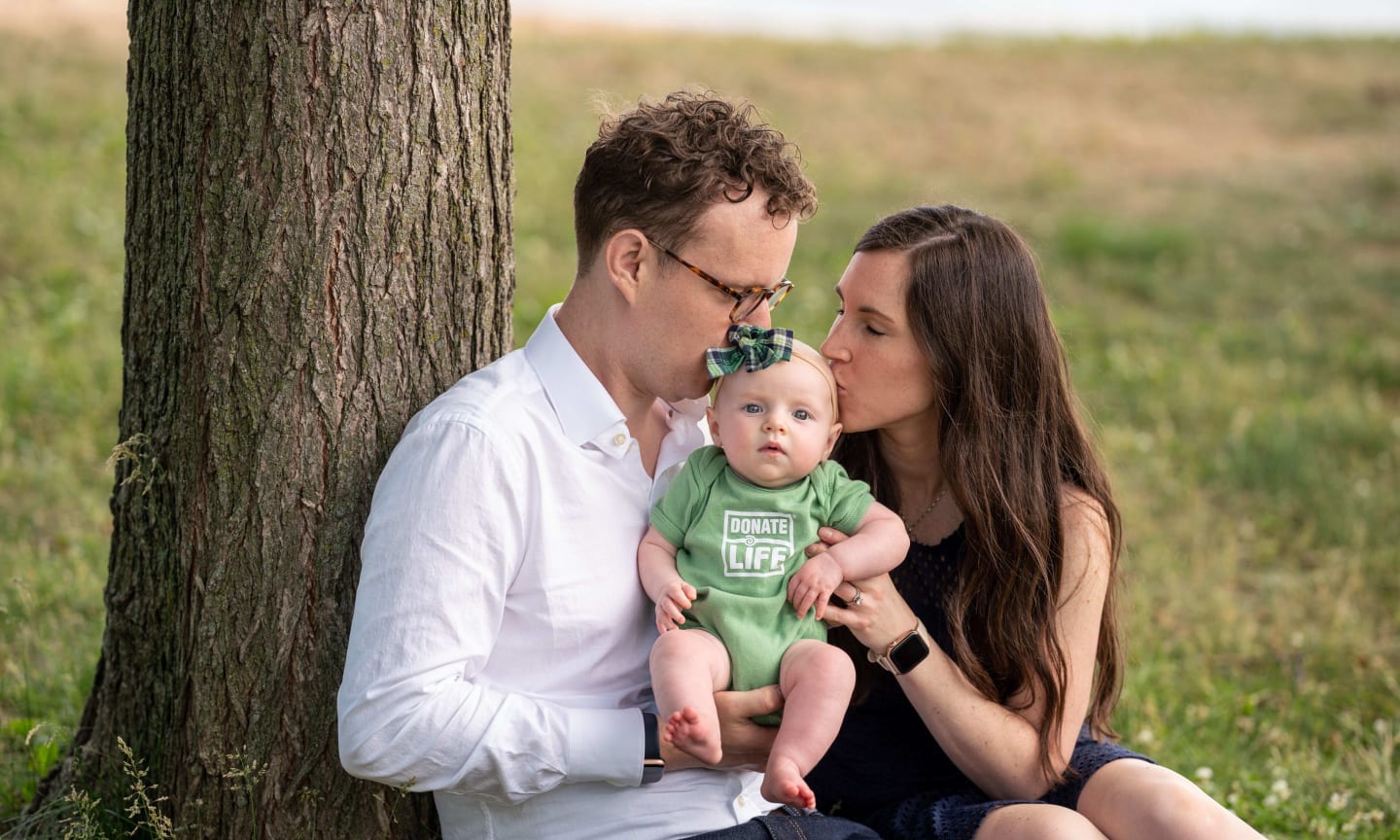 Nathan and Ann Bagget kissing their baby, Lucy, in front of a tree