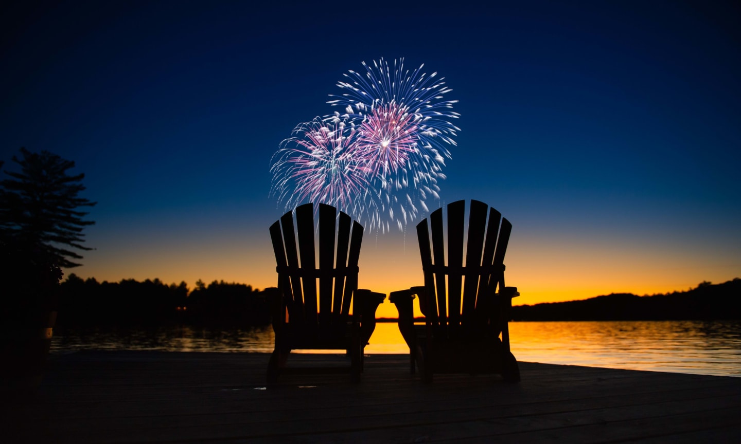 Canada day fireworks on a lake in Muskoka, Ontario Canada. On the wooden dock two Adirondack chairs are facing the sunset orange hues
