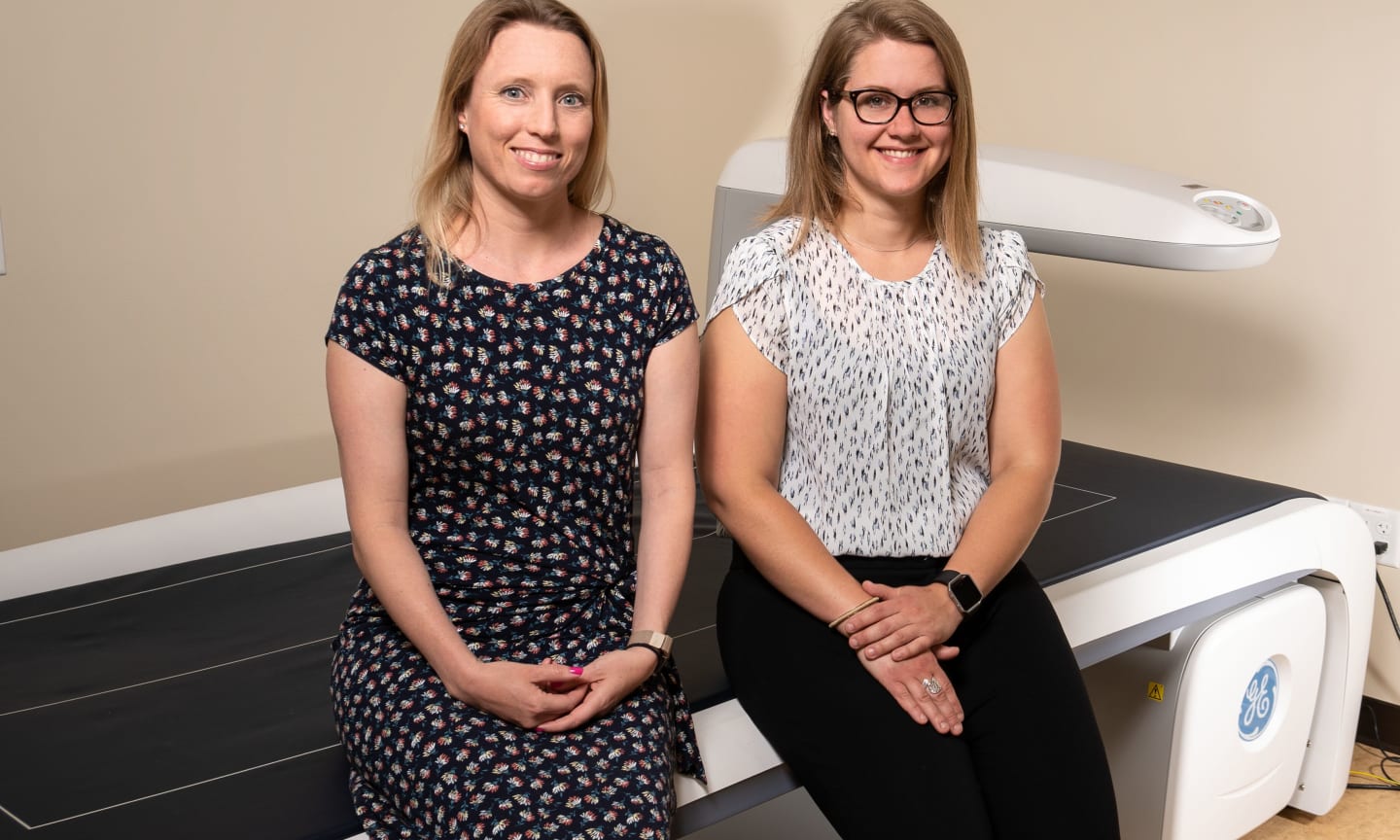 Lisa Cadmus-Bertram and Jess Gorzelitz sitting on a medical imaging machine