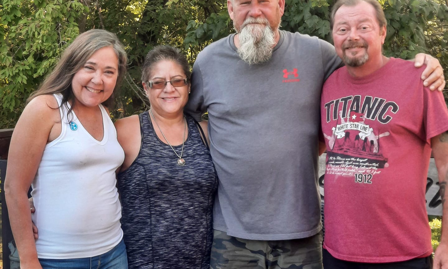 Four adults, two women and two men, facing the camera smiling with their arms around each other.