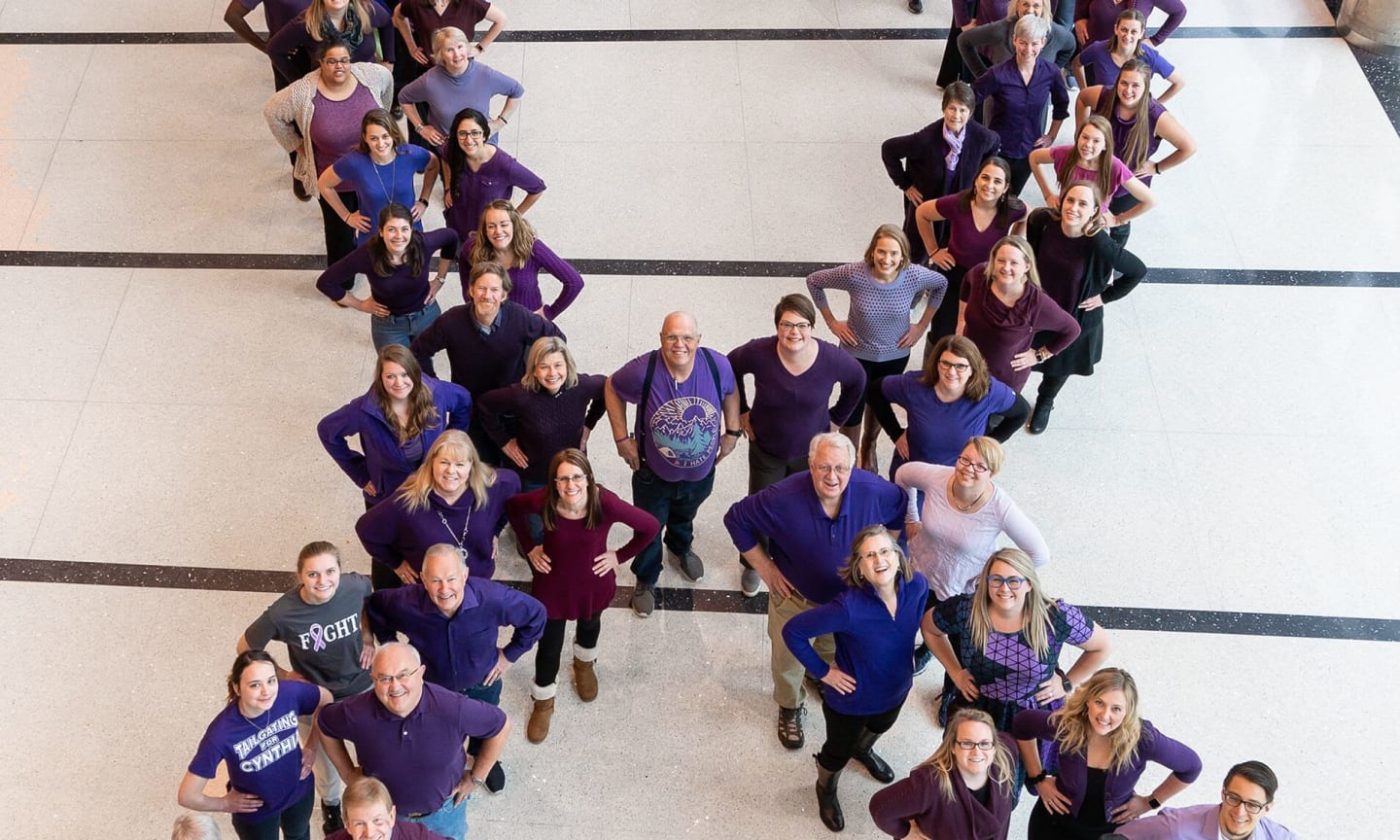 A group of people, all wearing purple, stand in the shape of a ribbon for pancreatic cancer