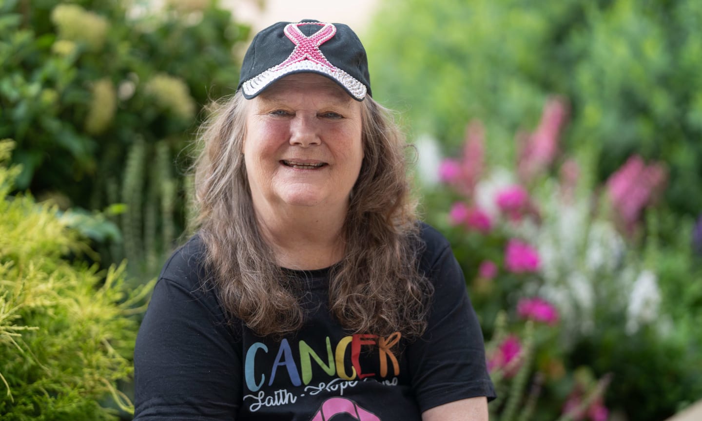 A woman wearing a black hat and a black shirt with pink ribbons on them smiling and sitting in a garden