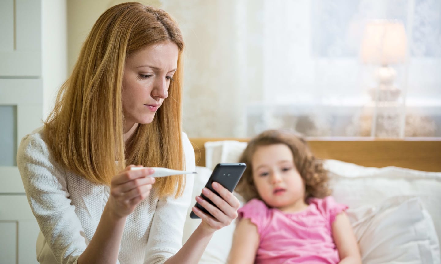 Mother measuring temperature of her ill kid. Sick child with high fever laying in bed and mother holding thermometer. Mother with cell phone calling to doctor