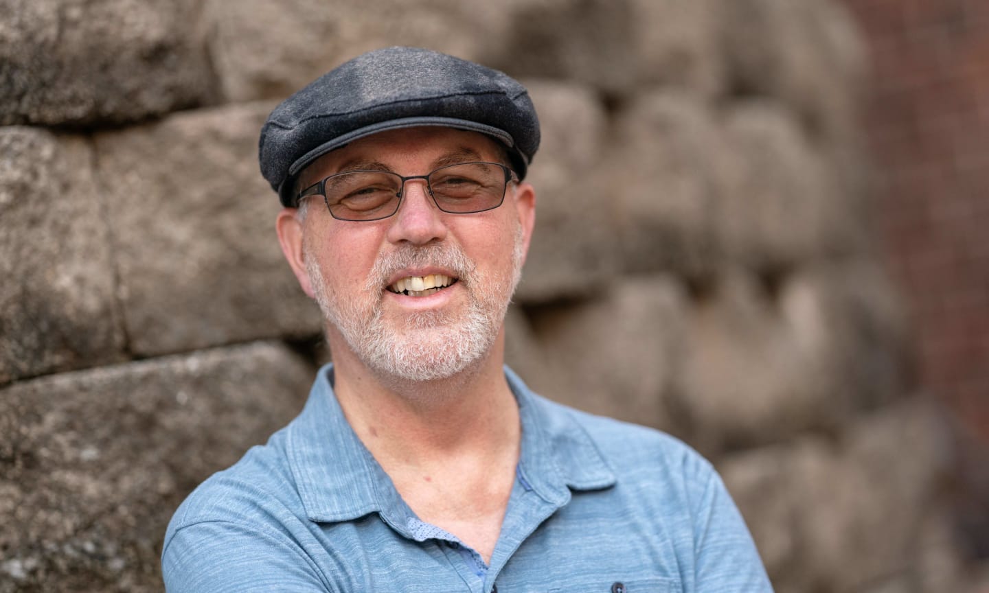 Randy Hatfield, dressed in a blue collared shirt and wearing glasses and a black hat, smiling outside.