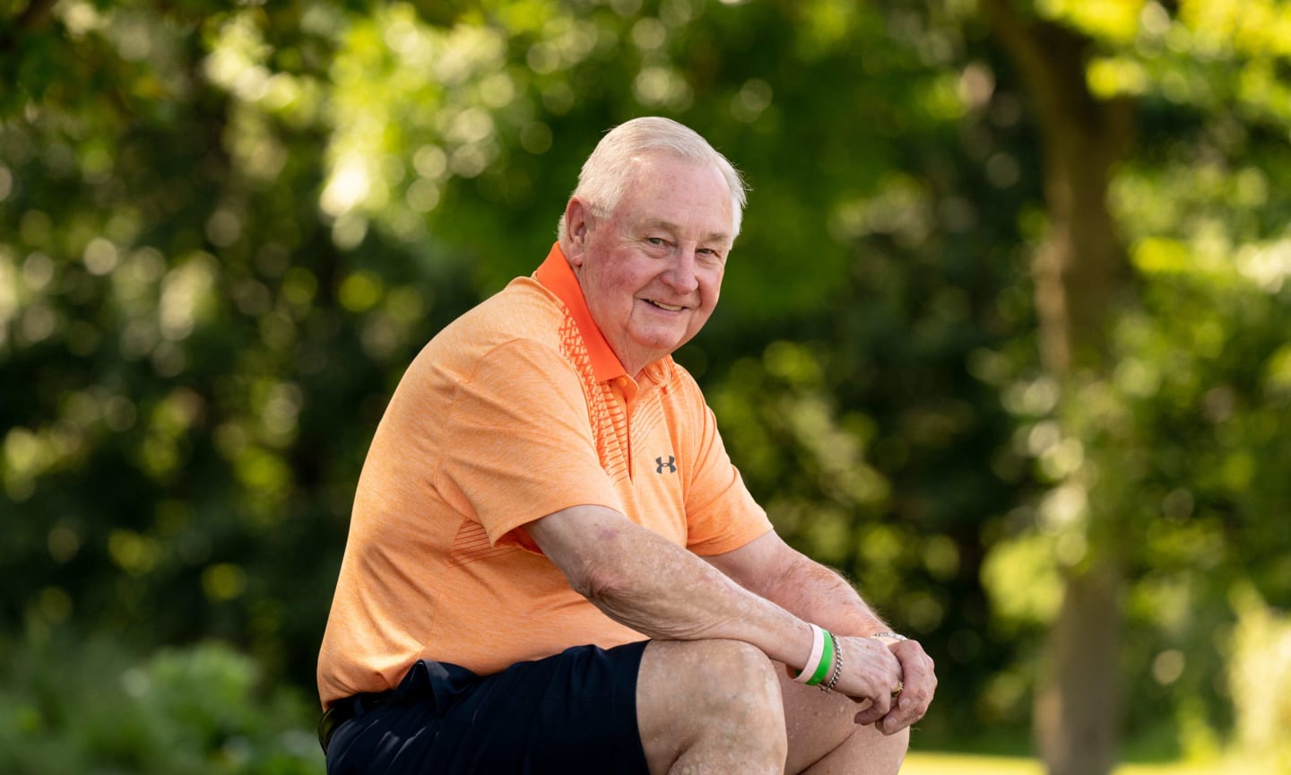 Bill Lawton, wearing an orange polo shirt and black shorts, seated outside on a sunny day