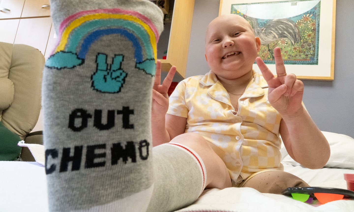 Girl sitting up in bed, smiling, with bottom of foot in foreground, with a sock on that says, "Peace out, chemo."