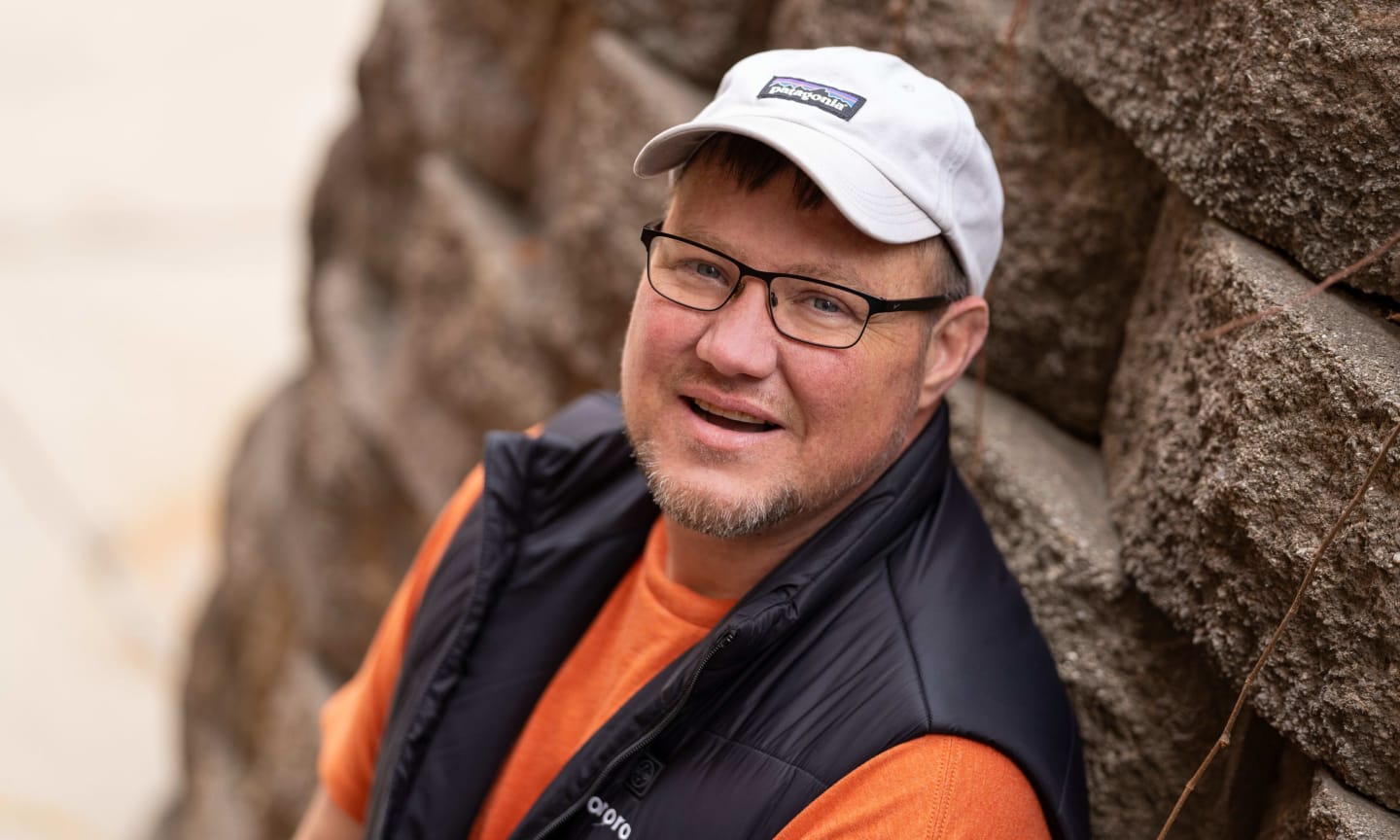 A man in a shite cap and glasses, smiling and leaning against a stone wall.