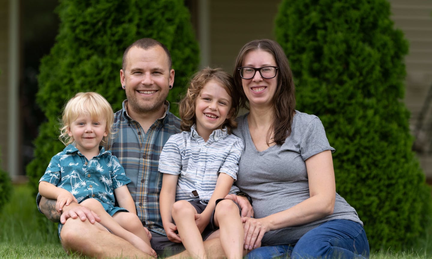 A mother and a father smiling, sitting in the grass outside, and holding two small boys on their laps.