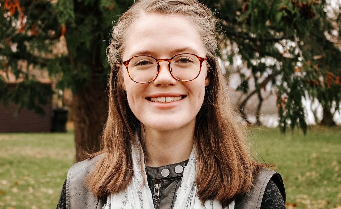 Young woman smiling outdoors in front of a tree, wearing glasses and a white scarf.