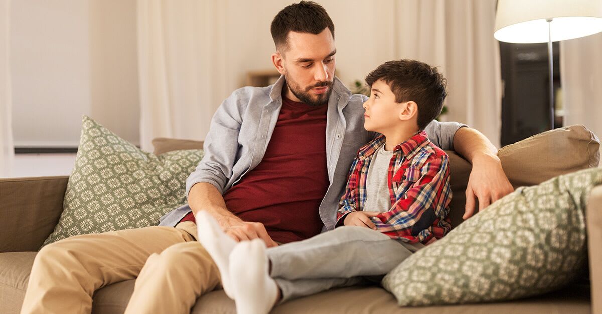 A father with his arm around his son, sitting on a couch