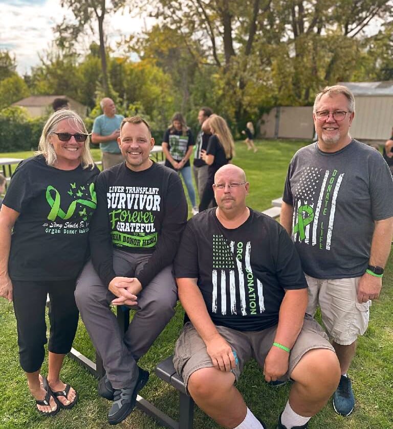 Four adults smiling outdoors, one female and three males.