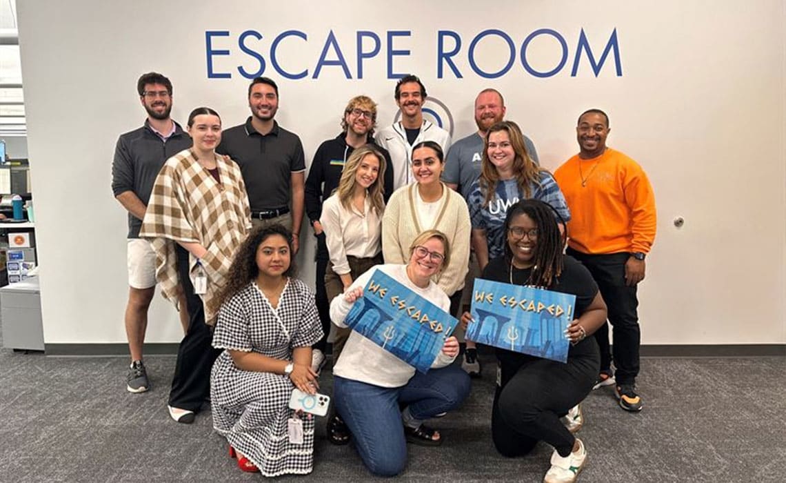 Diverse team happily posing in front of UWMs escape room with 'we escaped!' signs, after successfully collaborating as a team to solve the puzzles and escape.