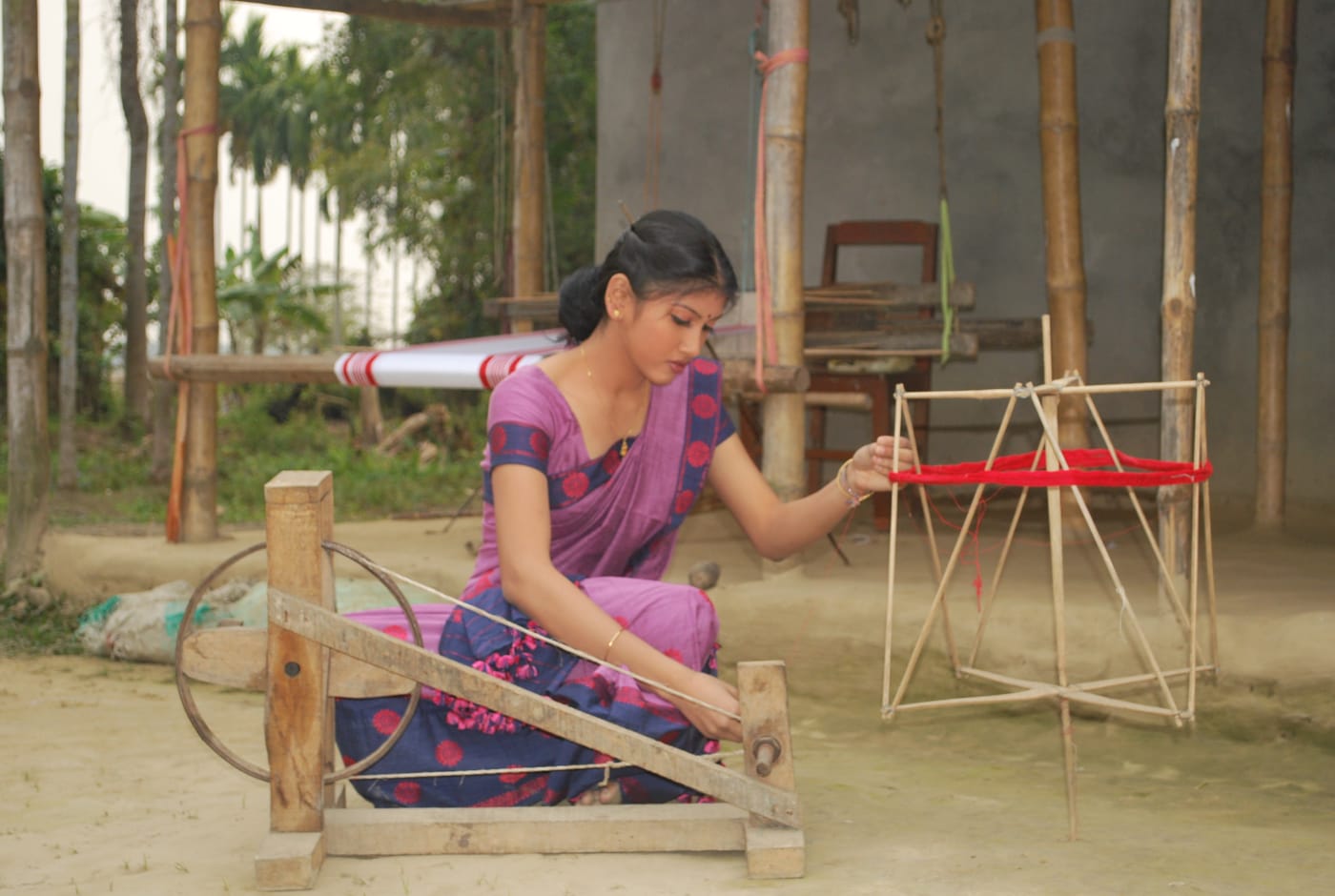 A spinner at her wheel in Assam, a loom behind her.