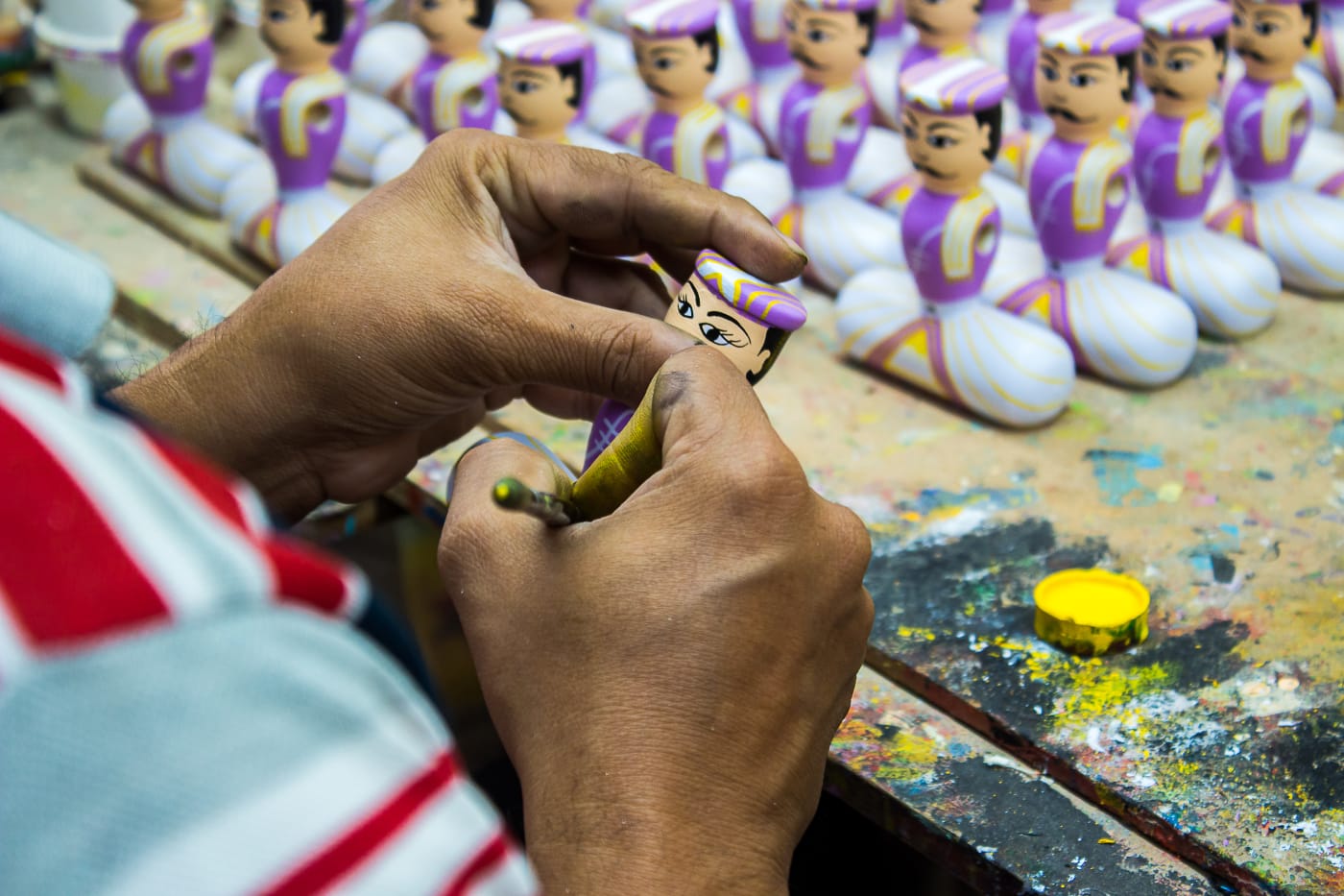 An artisan paints a wooden Channapatna doll, with rows of finished dolls behind, in Channapatna, Karnataka.
