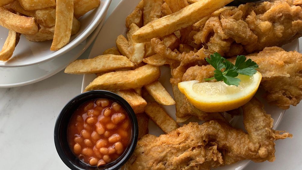 baked beans in small black bowl with potato chips with fried fish and lemon wedge and green leaf as garnish