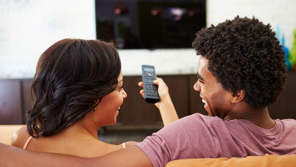 a man and a woman sitting on a sofa facing and smiling at each other with remote in woman's hand. there is television and t v console in the background