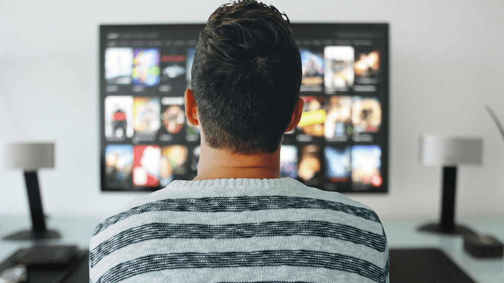 The back of a man's head watching Television, you can see him scrolling through a streaming service on the screen