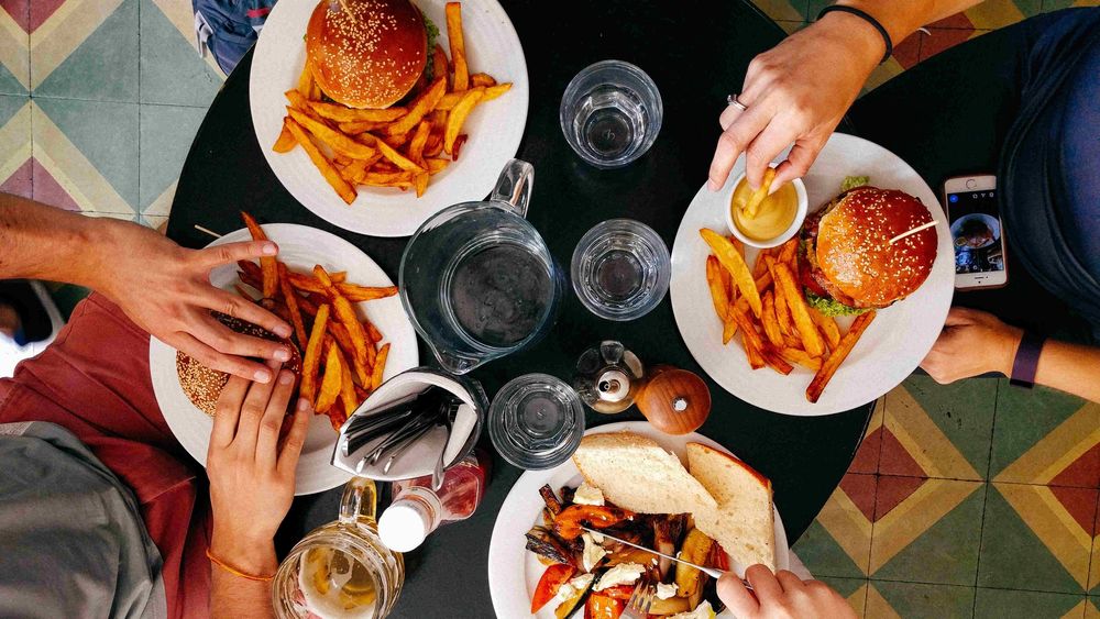 four white plates of burgers and chips with set of hand overing over three of them and clear glasses and jucg and salt shaker and bottle of ketchup