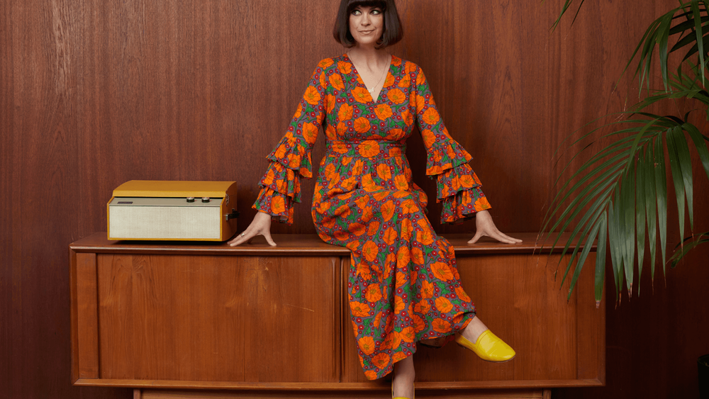 Women in floral dress sitting on cupboard next to a tree with a wooden wall behind her