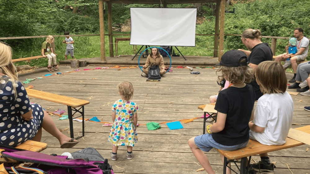 women holding hula hoop surrounded by children on wooden benches in a circular shape with a forest in the background