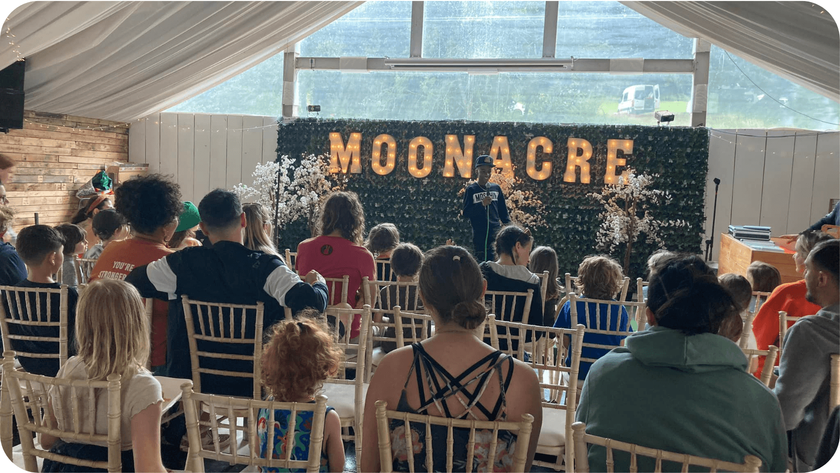 people sitting on chairs in a tent watching a boy do a speech on a stage