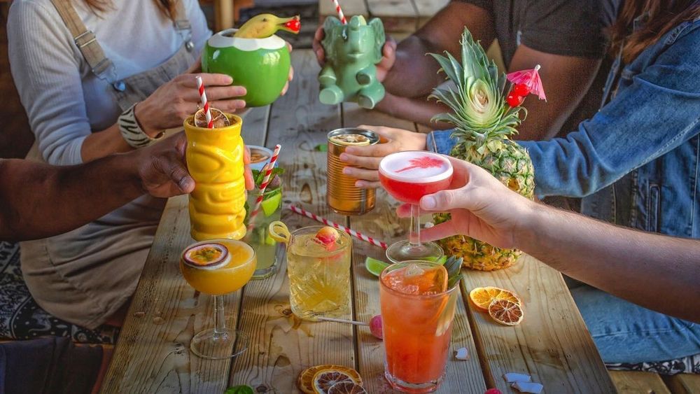 Group of people sat round an outside wooden bench doing a cheers with a variety of different coloured cocktails