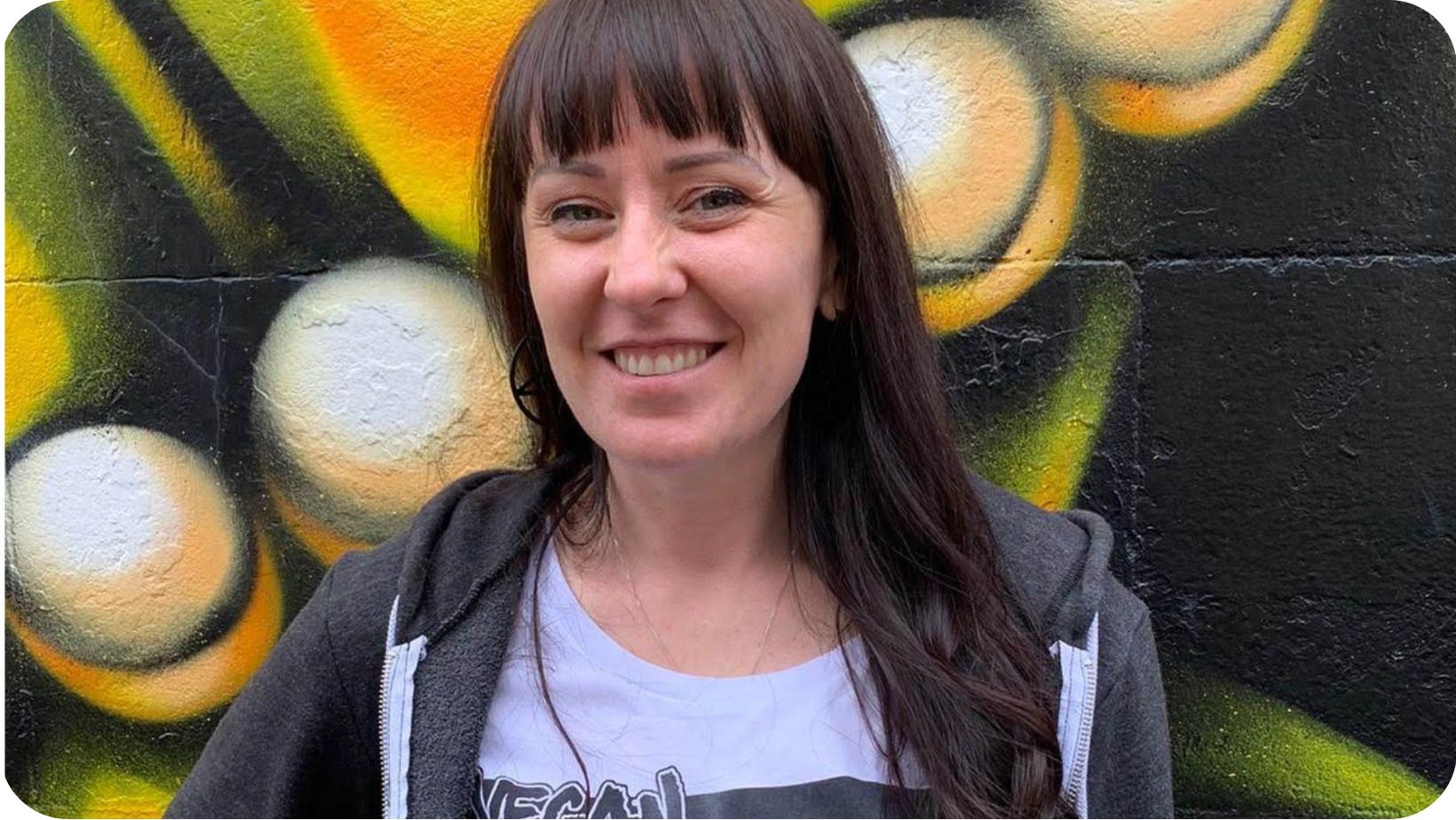 smiling woman with long, dark hair with a multicoloured graffiti wall in the background