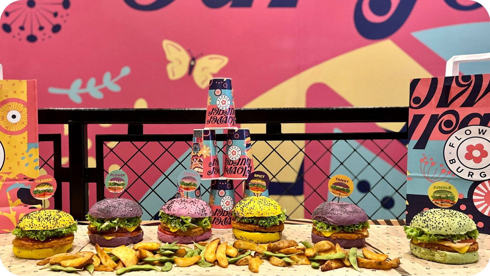 six multicoloured vegan burgers with potato wedges on a table in front of a multicoloured graphic-based wall
