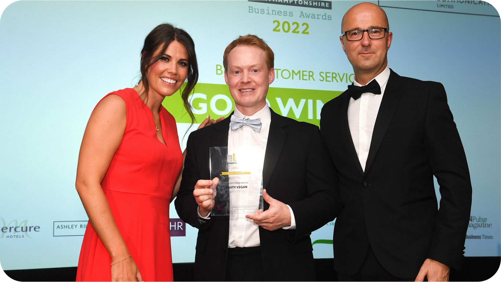 man in a suit and tie smiling accepting an award next to a smiling woman in a red dress and a man with a suit 