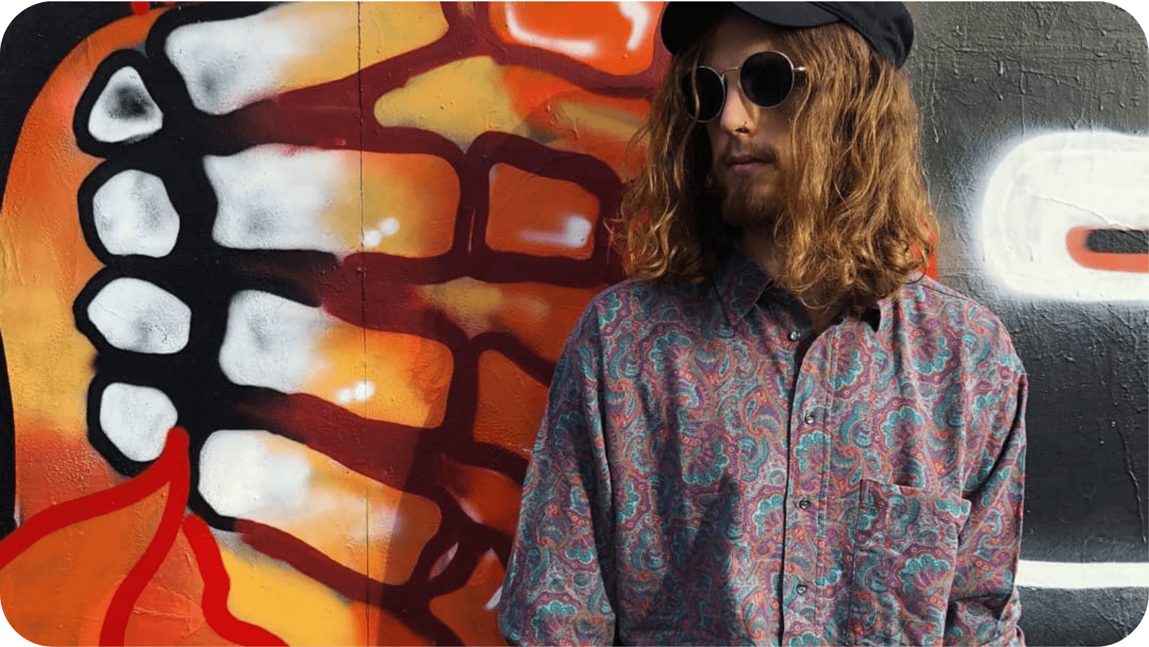 Man with long hair, sunglasses and hat with a multicolour print shirt in front of a wall with graffiti