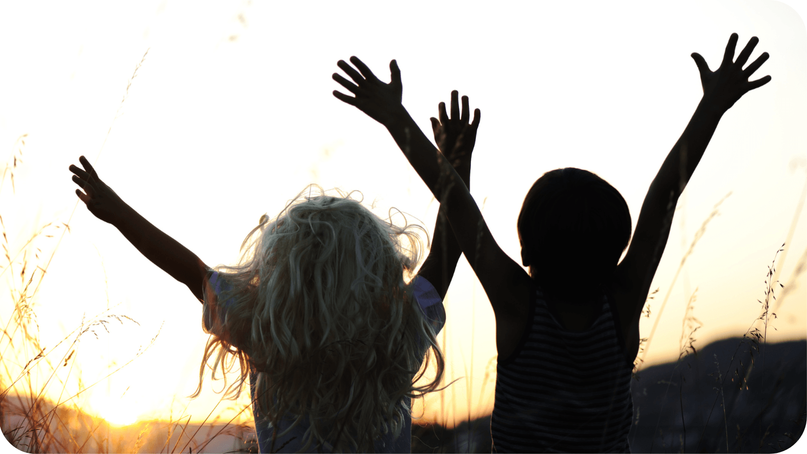 two children joyfully putting their hands in their with a backdrop of the setting sun in a field