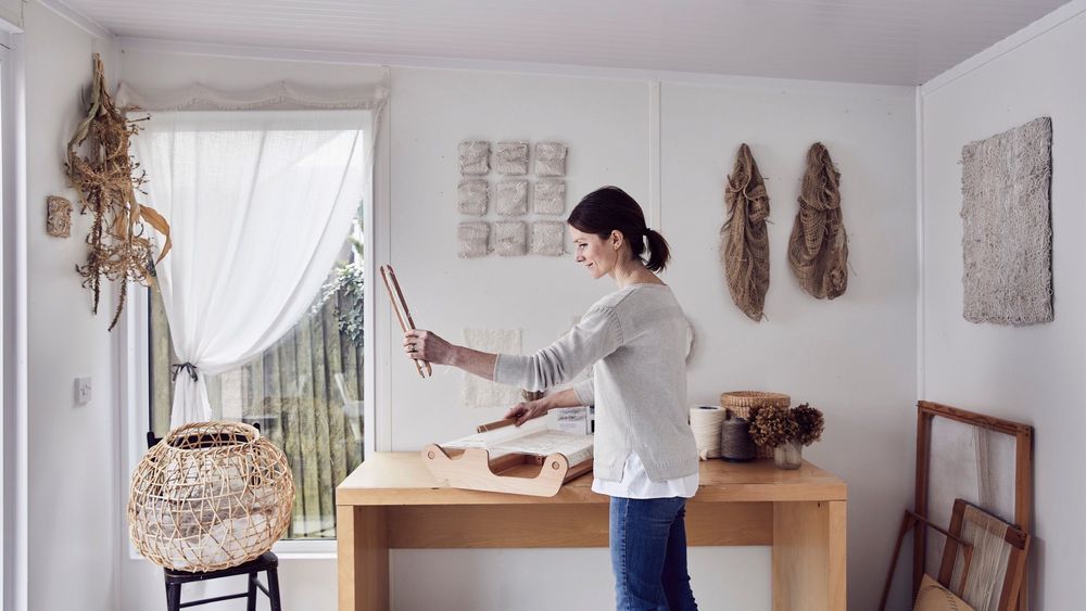 artist holding brushes in her art studio surrounded by a variety of pieces such as paintings and weaved baskets