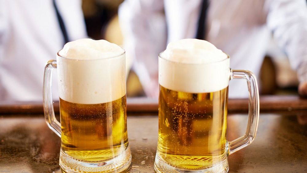 two pints of beer in glass on a wooden counter with two men chatting in the background