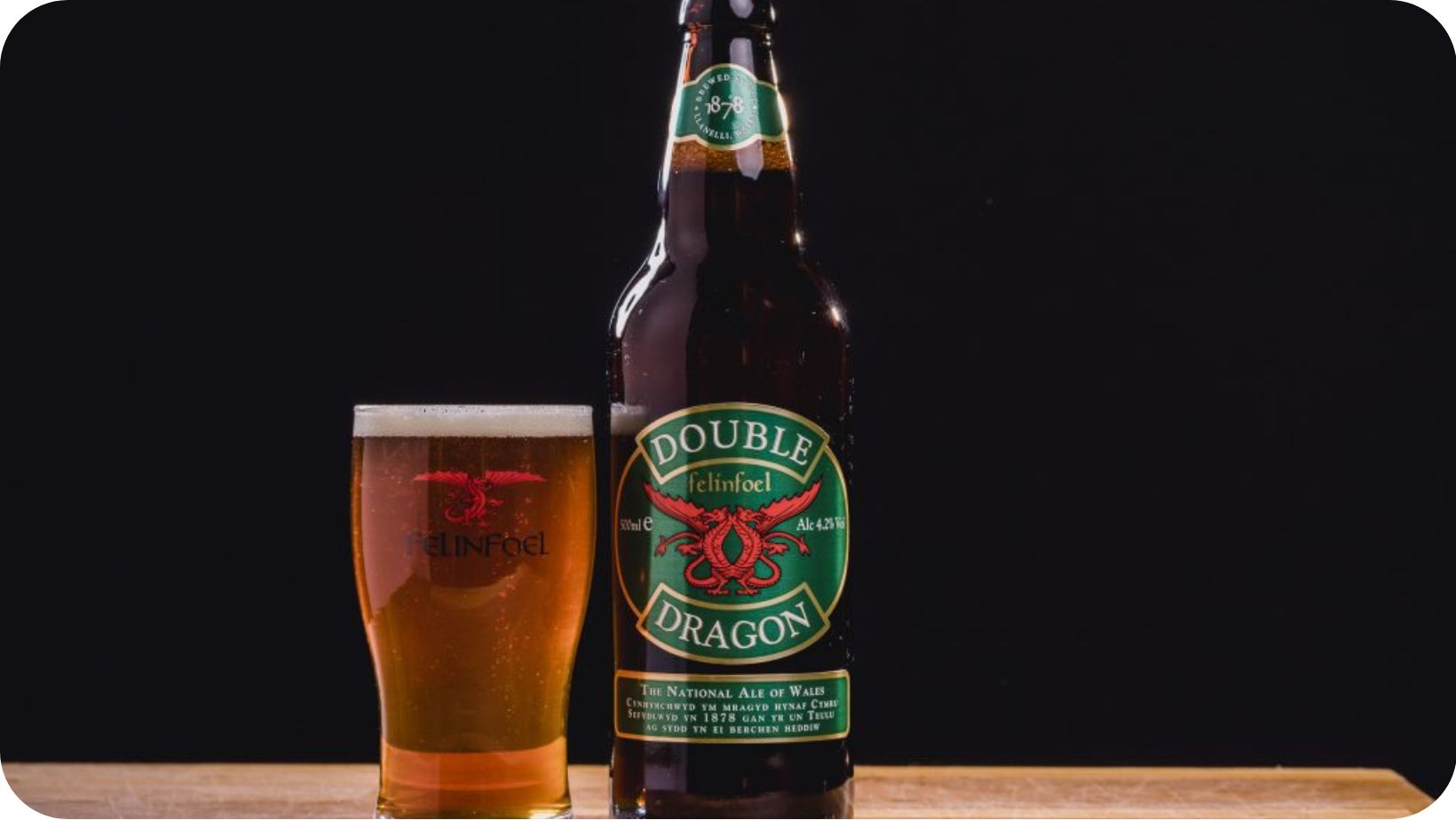 Bottle of Double Dragon vegan ale on a wooden table with a black background behind next to a full pint glass full of the ale