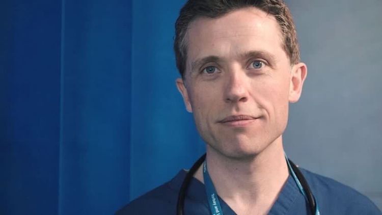 Male Consultant Gastroenterologist and General Physician (Dr Alan Desmond) smiling with a blue medical top on in front of a blue background