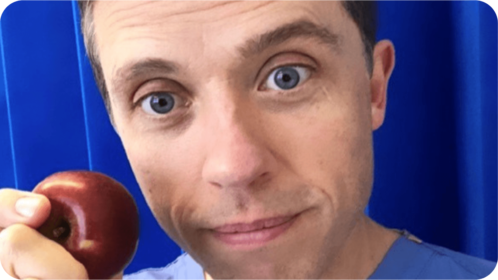 Male Consultant Gastroenterologist and General Physician (Dr Alan Desmond) smiling and a holding a red apple with a blue medical top on in front of a blue background