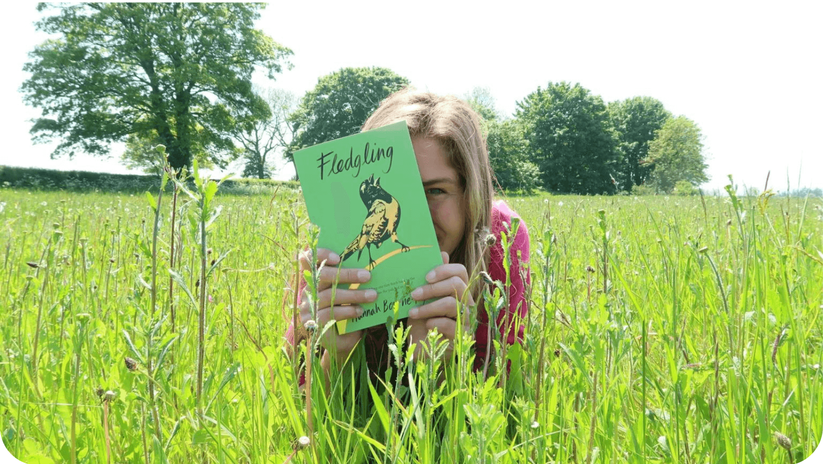 Woman (Hannah Bourne-Taylor) lying down in a sunny field of long grass hiding her face behind her book Fledging