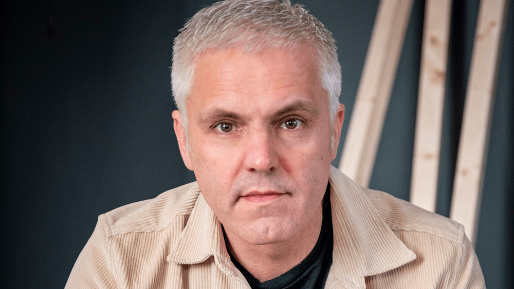 Man (Matthew Glover) with short grey hair smiling, wearing a beige coat in front of a black background 