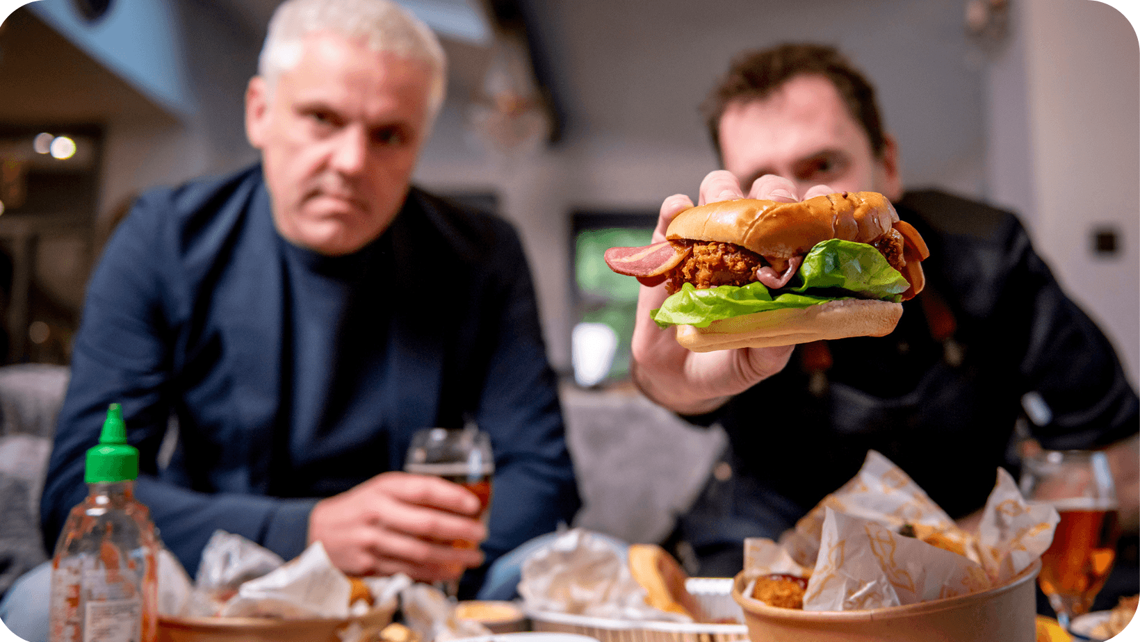 Two men: one holding a vegan burger and the other (Matthew Glover) holding a glass of red wine - they’re sitting down behind a table full of vegan food